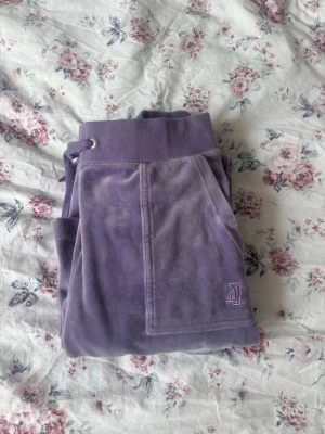 Lila velourbyxor Juicy Couture - Säljer ett par lila mjukisbyxor från Juicy Couture i mjuk velour. Byxorna har bred resår i midjan med dragsko, fickor fram och den klassiska Juicy-loggan broderad på ena benet. Perfekta för en chill och trendig look.
