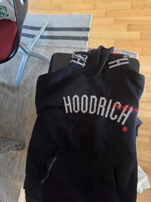 Svart HOODRICH hoodie med tryck - Snygg svart hoodie från HOODRICH med stor vit logga framtill och coolt tryck på ryggen. Huva med grå detaljer och känguruficka framtill. Perfekt för dig som gillar streetwear och vill sticka ut med en unik design. Passar alla som är mellan 170-183