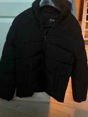 Svart pufferjacka från Only & Sons - Snygg svart pufferjacka från Only & Sons med dragkedja framtill och hög krage. Jackan har en klassisk quiltad design och är perfekt för kalla dagar. Tillverkad i ett slitstarkt material som håller värmen bra. Priset är ej hugget i sten. Skriv för frågor. Sitter slim