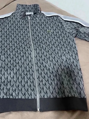 Mönstrad zip hoodie från Lacoste - Snygg grå och svart zip hoodie från Lacoste med ett geometriskt mönster och vit rand längs ärmarna. Klassisk krokodil-logga på bröstet och hög krage. Tillverkad i mjukt material som känns bekvämt mot huden. Perfekt för en sportig och trendig look.