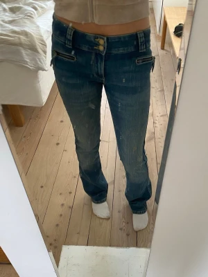 Blå bootcut jeans med dubbla knappar - Snygga blå bootcut jeans med dubbla knappar i midjan och dragkedjor vid fickorna framtill. Jeansen har en klassisk tvätt och raka ben med lätt utsvängd passform. Perfekta för en trendig och avslappnad look. 170