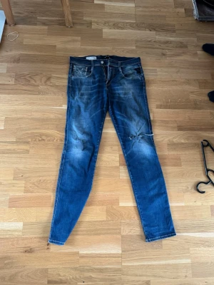 Blå slim fit jeans från Replay - Säljer ett par blå slim fit jeans från Replay med slitningar och detaljerade tvättade partier. Jeansen har klassisk femficksdesign, dragkedjegylf och snygga slitna effekter på benen. Perfekta för en avslappnad och trendig look. De är hyperflex och passar som en smäck
