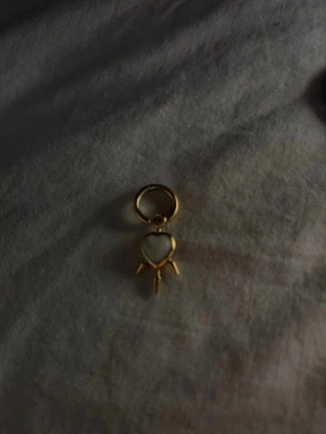 Maria Nilsdotter Guldigt örhänge med hjärthänge - Unikt örhänge i guldfärgad metall med ett hängande hjärta. Ena sidan av hjärtat är vit och den andra är guldig. Hängsmycket har små dekorativa detaljer nedtill. Perfekt för dig som gillar trendiga och lekfulla accessoarer. Super fin och använda inte sååå många gånger.
