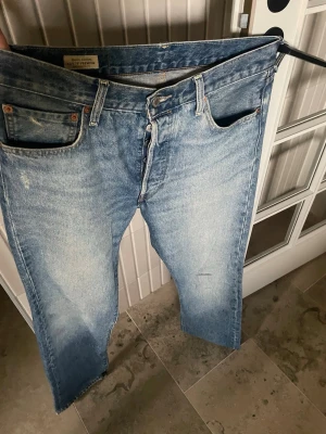 Levi's 501 ljusblå raka jeans - Klassiska Levi's 501 jeans i ljusblå denim med raka ben och femficksmodell. Jeansen har en vintage tvätt och subtila slitningar för en avslappnad look. Tillverkade i bomull och har knappgylf samt ikonisk läderpatch bak.