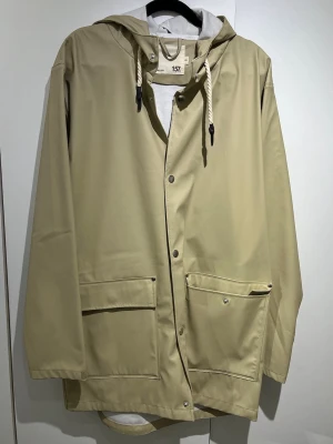 Beige regnjacka med huva - lager 157 - Stilren beige regnjacka från 157 med huva och vita dragsnören. Jackan har tryckknappar framtill, två stora fickor med lock och en rak passform. Materialet är vattenavvisande och perfekt för regniga dagar. Enkel och modern design som passar till det mesta.