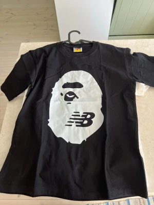 Svart BAPE x New Balance t-shirt - Säljer en svart t-shirt med stor vit BAPE-apa och New Balance-logga på bröstet. Klassisk rund hals och korta ärmar. Perfekt för dig som gillar streetwear och vill sticka ut med en unik collab.