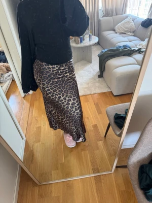 Leopardmönstrad långkjol i satin - Snygg långkjol med leopardmönster i bruna och svarta toner. Kjolen har en rak passform och är gjord i ett glansigt satinliknande material som ger en lyxig känsla. Perfekt för dig som vill sticka ut med ett djurmönster. Aldrig använd 