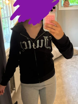 Svart Juicy Couture hoodie med strass - Säljer en svart hoodie från Juicy Couture i mjuk velour med dragkedja framtill och huva. På bröstet står det 'Juicy' i stora bokstäver med glittrande strassdetaljer. Hoodien har långa ärmar och fickor framtill. Perfekt för en chill och trendig look.