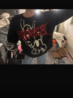 Svart stickad tröja med grafiskt tryck - Cool svart stickad tröja med stort vitt hundmotiv och röd graffiti-text på bröstet. Ena ärmen har zebramönster i svart och vitt. Oversized passform och rund hals. Perfekt för dig som gillar streetwear och unika plagg.