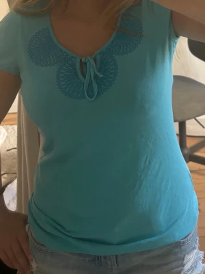 Turkos topp med knyt från Esprit - Snygg turkos topp från Esprit med korta ärmar och dekorativt knyt i halsen. Toppen har ett broderat mönster runt ringningen som ger en unik touch. Perfekt till jeans eller shorts för en fräsch look.