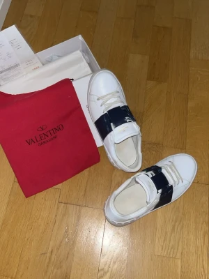 Valentino Garavani vita sneakers - Säljer ett par vita sneakers från Valentino Garavani med bred marinblåa läderdetalj över snörningen och subtila nitar på hälen. Skorna har en klassisk låg profil och är tillverkade i slätt läder. Perfekta för dig som gillar exklusiv och stilren design.