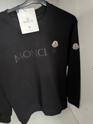 Svart Moncler sweatshirt med logga - Cool svart sweatshirt från Moncler med diskret logotyp på bröstet och ärmen. Tröjan har rund halsringning och är tillverkad i mjukt material som passar perfekt för en avslappnad stil. Moncler-märke på både bröst och ärm ger en exklusiv känsla.
