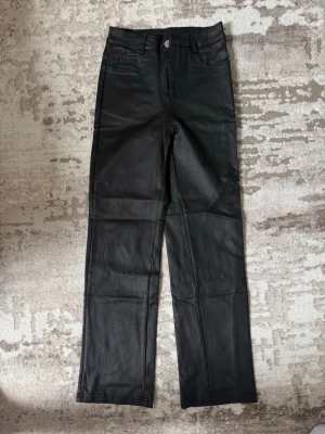 Svarta coated raka jeans - Säljer ett par svarta coated jeans med rak passform och hög midja i petite. Jeansen har klassiska fem fickor och stängs med knapp och dragkedja. Materialet har en lätt glansig finish som ger en cool look.