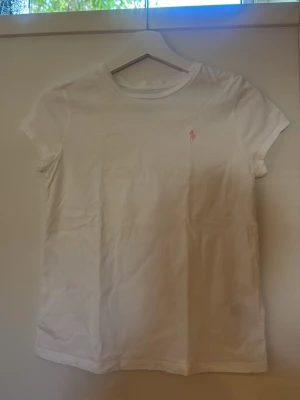Vit t-shirt från Polo Ralph Lauren - Klassisk vit t-shirt från Polo Ralph Lauren med korta ärmar och rund hals. T-shirten har en liten rosa broderad logga på bröstet och är gjord i mjuk bomull. Perfekt basplagg för en clean och enkel stil.