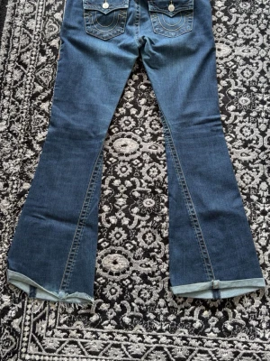 Blå bootcut jeans från True Religion - Säljer ett par blå bootcut jeans från True Religion med klassiska kontrastsömmar och stora fickor bak. Jeansen har råa, fransiga benslut och en snygg utsvängd passform. Perfekta för dig som gillar retrovibbar och streetstyle.