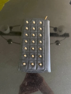 Svart plånbok med nitar - Svart plånbok i konstläder med guldfärgade nitar på framsidan. Plånboken har dragkedja runt och en stilren, rektangulär form.