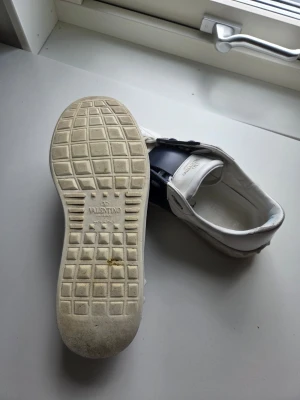 Valentino vita sneakers med blå detalj - Snygga vita sneakers från Valentino med bred mörkblå rem över sidan. Skorna har klassisk låg profil, vita snören och diskret logga på sulan och plösen. Tillverkade i läder med gummisula. Perfekta för dig som gillar stilrena och exklusiva sneakers.