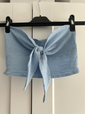 Ljusblå bandeau topp med knyt - En ljusblå bandeau topp med knyt framtill. Toppen är ärmlös och har en smockad, stretchig struktur som sitter bekvämt. Perfekt för varma dagar och enkel att styla med jeans eller kjol.
