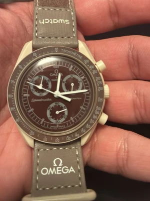 Omega x Swatch Speedmaster Mocha - Snygg Omega x Swatch Speedmaster Mocha med brun urtavla och beige boett. Klockan har kronograf, tachymeter och tydliga vita visare. Matchande beige textilband med Omega och Swatch-logga. Perfekt för dig som gillar stilrena accessoarer.