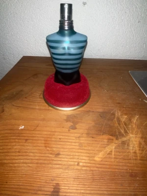 Jean Paul Gaultier Le Male parfym sampels 3 ml - Ikonisk parfymflaska formad som en manlig torso i blått glas med turkosa och mörkblå ränder. Flaskan har metallock och står på en rund röd sammetsliknande bas. En klassiker från Jean Paul Gaultier som sticker ut i badrumshyllan.