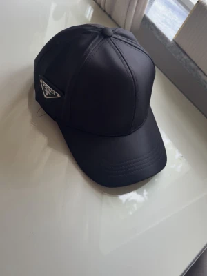 Svart keps från Prada i nylon - Snygg svart keps från Prada i glansigt nylonmaterial med klassisk böjd skärm och ikonisk metallogo på sidan. Perfekt accessoar för en trendig och stilren look. Utan prada lapp 