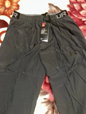 Svarta mjukisbyxor från Under Armour - Snygga svarta mjukisbyxor från Under Armour med elastisk midja och dragsko. Byxorna har diskret logga på benet och dragkedjeförsedda fickor. Tillverkade i ett lätt och mjukt material som känns skönt mot huden. Perfekta för chill eller träning.