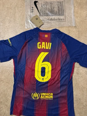 FC Barcelona matchtröja Gavi #6 - Säljer en officiell FC Barcelona matchtröja från Nike med Gavi och nummer 6 på ryggen. Tröjan är blå och röd med gula detaljer, klubbmärke och sponsorer som Spotify och UNHCR. Tillverkad i lätt och ventilerande material, perfekt för fotboll eller träning.