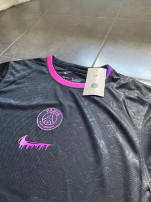 Svart PSG x Nike t-shirt med lila detaljer - Snygg svart t-shirt från Nike i samarbete med Paris Saint-Germain. Tröjan har lila detaljer vid krage och ärmslut, PSG-logga på bröstet och en unik lila Nike swoosh med drip-effekt. Tillverkad i ett lätt och ventilerande material, perfekt för träning eller streetwear.