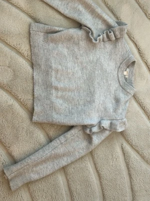 Ljusgrå stickad tröja med volang - Supersöt ljusgrå stickad tröja med volangdetaljer på axlarna. Tröjan har rund halsringning, långa ärmar och ribbade muddar vid ärmslut och nederkant. Perfekt för dig som gillar en enkel men trendig look.