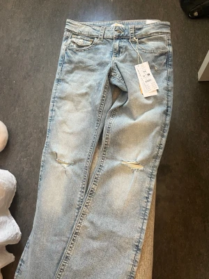 Ljusblå Lågmidjade slitna jeans  - Säljer ett par ljusblå jeans från Zara med raka ben och slitningar vid knäna. Jeansen har klassisk femficksdesign och är gjorda i denim med en avslappnad passform. Perfekta för en trendig och avslappnad stil. St 164