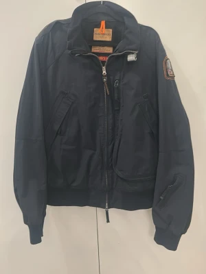 Svart vindjacka från Parajumpers - Stilren svart vindjacka från Parajumpers med flera fickor, hög krage och orange detalj i nacken. Jackan har patch på ärmen och robust dragkedja framtill. Perfekt för dig som vill ha en sportig och urban look.