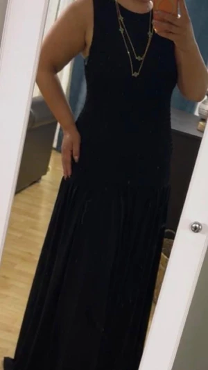 Svart långklänning på med ärmlös design - Stilren svart långklänning med ärmlös design och rund halsringning. Klänningen har en smockad midja och ett mjukt, fallande tyg som ger en elegant siluett. Perfekt för dig som gillar enkel och tidlös stil. Köpt ifrån hm och inte använd alls! Modellen är storlek L men klänningen är stretchig.