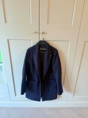 Mörkblå ullrock från Herno - Snygg mörkblå rock från Herno med klassisk krage och knäppning framtill. Rocken har två fickor på sidorna och är tillverkad i ett ullmaterial som håller dig varm. Perfekt för dig som vill ha en stilren och tidlös look. Nypris 9000kr.