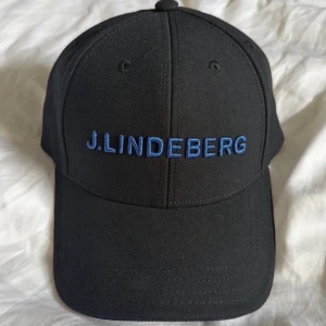 Svart keps från J.Lindeberg GE PRIS FÖRSLAG - Snygg svart keps från J.Lindeberg med böjd skärm och blå broderad logga framtill. Passar perfekt till golfrundan eller bara allmänt i vardagen. 