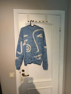Blå stickad tröja med ansiktsmotiv - Säljer en blåmelerad stickad tröja med vitt abstrakt ansiktsmotiv på framsidan. Tröjan har rund halsringning, långa ärmar och ribbade muddar. Perfekt för dig som gillar unik design och konstnärliga detaljer.