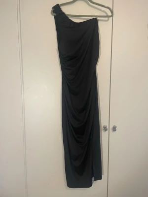 Svart långklänning  - Stilren svart långklänning med oneshoulder-design och draperad effekt längs framsidan. Klänningen har en slits på sidan för extra rörelsefrihet och en elegant siluett. Perfekt för dig som vill ha en klassisk men modern look.
