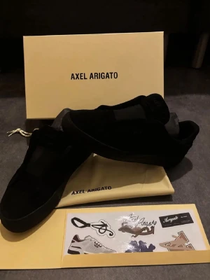 Svarta slip-on sneakers Axel Arigato - Säljer ett par stilrena slip-on sneakers från Axel Arigato i svart mocka. Minimalistisk design med elastiska band över vristen som gör dem enkla att ta på och av.  Skorna är i fint skick och kommer med: * Originalkartong * Dustbag * Originalstickers  Perfekta för dig som gillar en clean och exklusiv stil. Öppen för byten mot andra sneakers/skor!