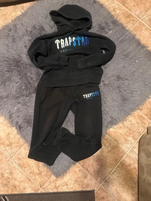 Trapstar set - Svarta mjukisbyxor och hoodie från Trapstar med broderad logga i vitt och blått på ena benet. Byxorna har resår i midjan och vid benslut samt sidofickor. Perfekta för en avslappnad och trendig streetwear-look.
