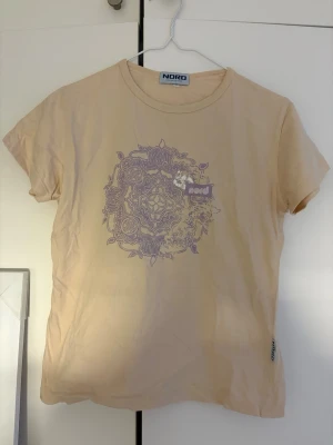 Beige t-shirt med lila tryck från NORD - Beige t-shirt från NORD med ett stort lila mandalainspirerat tryck på bröstet. Klassisk rund halsringning och korta ärmar. Tillverkad i mjuk bomull för en skön känsla. Liten NORD-tag på sidan ger extra detalj.