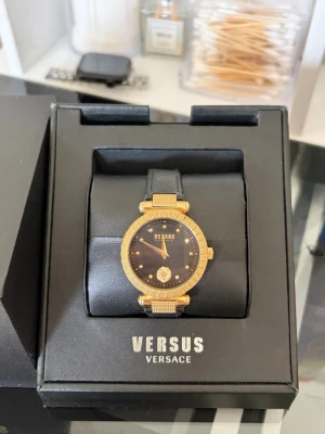 Svart och guld klocka Versus Versace - Snygg klocka från Versus Versace med svart urtavla och guldfärgad boett. Klockan har svarta band och detaljer i guld runt urtavlan samt Versus-logga. Perfekt accessoar för dig som gillar lyxig och stilren design.