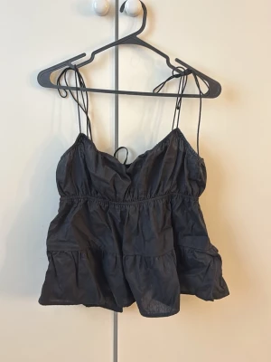 Svart sommartopp - Svart sommartopp från H&M med tunna axelband som knyts på axlarna. Toppen har en luftig och voluminös passform med rynkade detaljer och är tillverkad i mjuk bomull. Perfekt för varma dagar och enkel att styla med olika outfits. Jättefin till sommaren! Gåva vid köp!