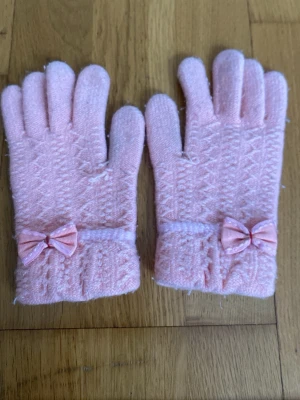 Rosa stickade handskar med rosett för barn - Söta rosa stickade handskar med dekorativa små rosetter på mudden. Handskarna har ett diskret mönster och är perfekta för kyliga dagar. De är mjuka och bekväma, och ger en gullig touch till din outfit.
