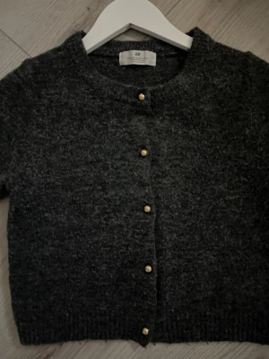 Mörkgrå kofta från H&M - Mysig mörkgrå kofta från H&M med rund halsringning och guldfärgade knappar framtill. Kortare modell med ribbade muddar vid ärmslut och nederkant. Perfekt att slänga över en topp för en chill look.