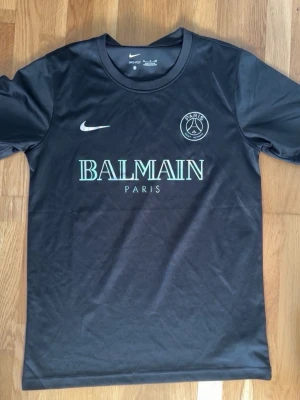 Svart PSG x Balmain t-shirt - Snygg svart t-shirt från Nike i samarbete med PSG och Balmain. Tröjan har rund hals, korta ärmar och tryck med Balmain Paris-logga samt PSG-emblem på bröstet. Tillverkad i ett mjukt och funktionellt material som passar perfekt till träning eller streetwear.