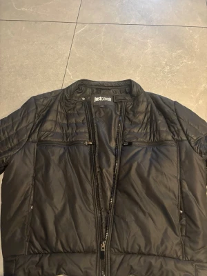 Svart pufferjacka från Just Cavalli - Säljer en svart pufferjacka från Just Cavalli med hög krage och dragkedja framtill. Jackan har två bröstfickor med dragkedja och två sidofickor. Materialet är glansigt och quiltat, perfekt för kalla dagar. Snygg och modern design som sticker ut.