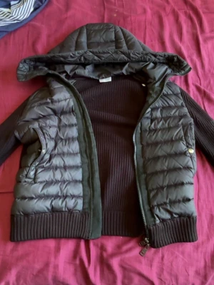 Svart moncler cardigan  - Snygg svart pufferjacka med stickade ärmar och rygg, samt vadderad framsida och huva. Jackan har dragkedja framtill, två fickor med knappstängning och ribbade muddar. Perfekt för kalla vinterdagar och ger en cool streetwear-look. Storlek S och pris kan sänkas vid snabb affär. ÄKTA