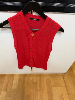 Röd stickad väst från Gina Tricot - Snygg röd stickad väst från Gina Tricot med v-ringad hals och guldfärgade knappar framtill. Västen har ett diskret hålmönster och ribbade kanter. 