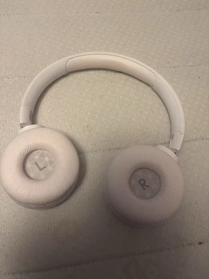 Vita on-ear hörlurar från JBL - Snygga vita on-ear hörlurar från JBL med vadderad bygel och mjuka öronkuddar. Hörlurarna har tydliga L och R-markeringar på insidan och en stilren design i plast och konstläder. Perfekta för musik, gaming eller videosamtal.
