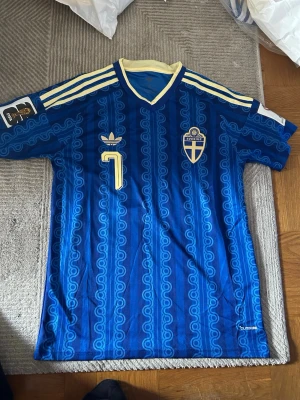 Blå Sverige retro fotbollströja Adidas - Retro fotbollströja från Adidas i blått med gula detaljer och nummer 7. Tröjan har Sveriges landslagsemblem på bröstet, klassiska Adidas-ränder på axlarna och ett unikt mönster i tyget. V-ringad krage i gult och blått. Perfekt för dig som älskar vintage sportstil.