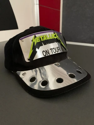 Y2K • Vintage • Metallica - ON TOUR Black Era • Snapback - Original vintage Metallica “ON TOUR” snapback-keps från sent 80-tal / tidigt 90-tal. Klassisk Metallica-relaterad vintage-keps från bandets storhetstid. Kepsen har den ikoniska neon-gröna Metallica-loggan monterad på en nitad metallplatta och formgjuten skärm – en design som är typisk för slutet av 80-talet och början av 90-talet och väldigt ovanlig.  En original vintage-produkt från perioden, inte någon nytillverkad replika eller modern kopia.